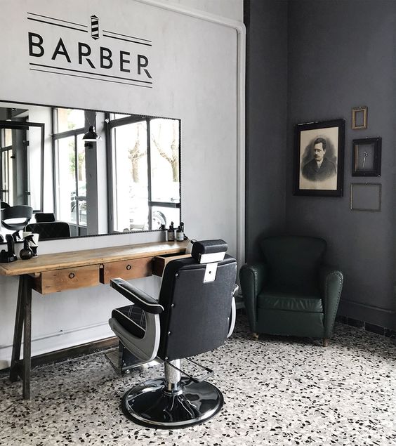 Rincón de una barbería. Una pared es blanca, la otra gris, el piso es blanco con manchas de colores. Hay un espejo en la pared, encima de un mueble, enfrente hay una silla y a su costado un sillon negro.