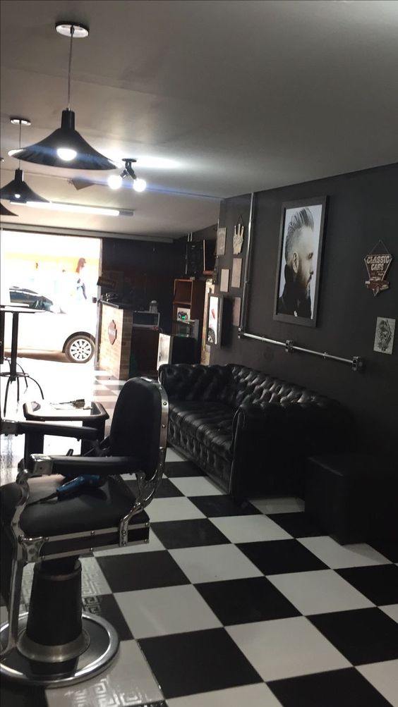 Foto del interior de una barbería. El techo es blanco  la pared negra y el piso ajedrezado en blanco y negro. La silla es negra, detras de ella se encuentra un sofá negro y una mesita en forma de cubo negra.